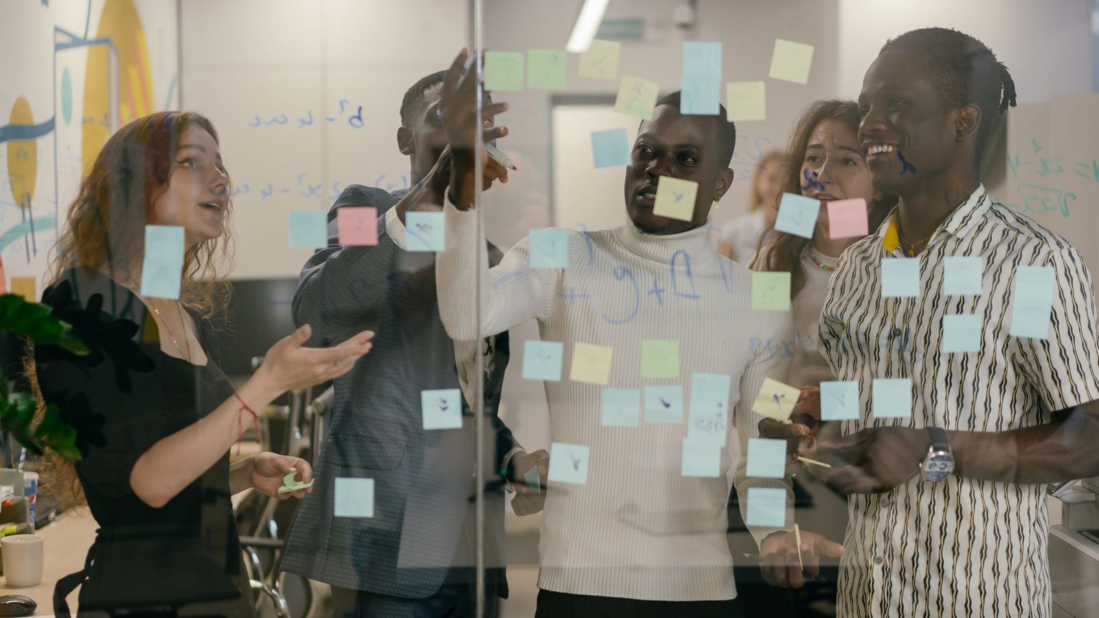 A diverse group of colleagues collaborates using sticky notes on a glass wall in an office